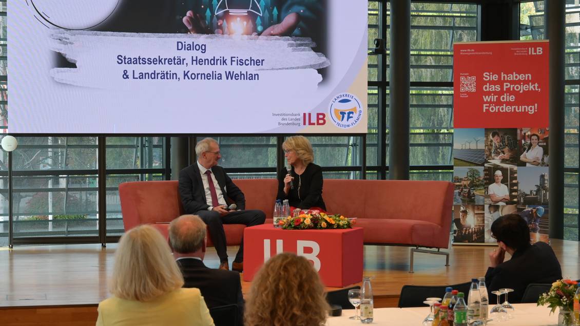 Das Foto zeigt zwei Personen im Kreistagssaal, die auf einer großen Couch sitzen und sich unterhalten. 