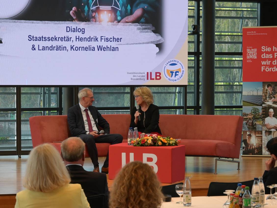 Das Foto zeigt zwei Personen im Kreistagssaal, die auf einer großen Couch sitzen und sich unterhalten. 