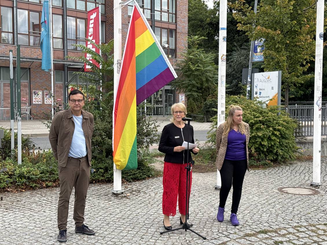 Das Foto zeigt drei Personen vor den Fahnenmasten vor dem Kreishaus in Luckenwalde. An einem Fahnenmast hängt eine Regenbogenflagge.