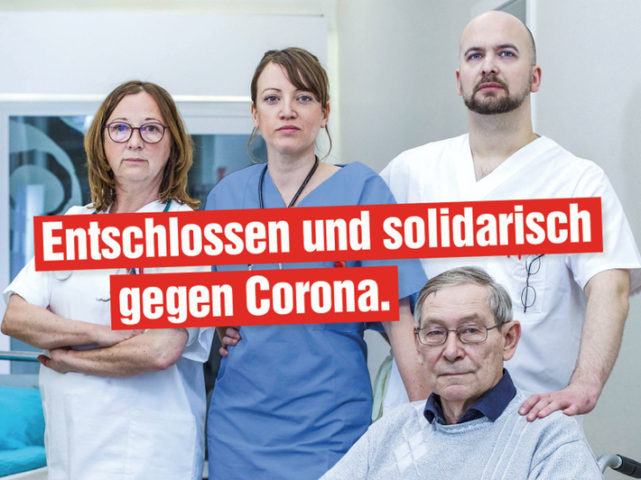 Das Foto zeigt vier Personen in einem Krankenhausflur, drei Personen sind Angestellte und stehen, eine Person ist Patient und sitzt. 
